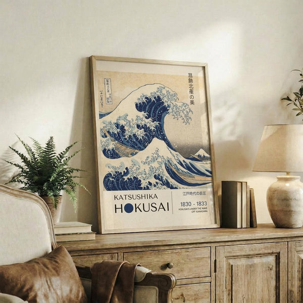May include: Framed poster of Hokusai's famous blue wave artwork with Japanese text and exhibition details, displayed on rustic wooden sideboard alongside fern plant, books, and ceramic vase in neutral-toned living room setting