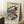 May include: Framed Japanese woodblock print artwork featuring a dramatic waterfall cascading over red-orange rocky cliffs with white foam and blue water, displayed in light wood frame leaning against white wall corner. Small wooden side table with white coffee cup visible. Traditional ukiyo-e art style with vibrant colors including yellows, greens, and blues depicting natural landscape scene.