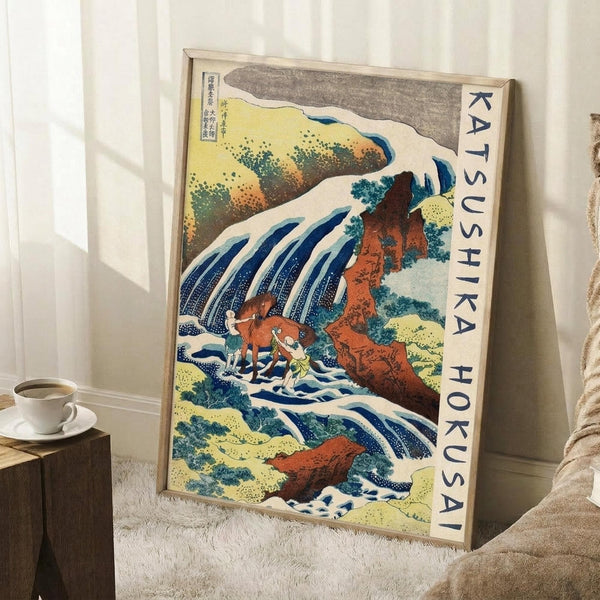 May include: Framed Japanese woodblock print artwork featuring a dramatic waterfall cascading over red-orange rocky cliffs with white foam and blue water, displayed in light wood frame leaning against white wall corner. Small wooden side table with white coffee cup visible. Traditional ukiyo-e art style with vibrant colors including yellows, greens, and blues depicting natural landscape scene.