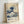 May include: Framed reproduction of Hokusai's famous Great Wave woodblock print displayed against white wall. The artwork shows a large blue wave with white foam threatening small boats, with Mount Fuji visible in background. Modern museum-style poster includes artist name "Katsushika Hokusai" and dates "1830-1833" in contemporary typography below the traditional Japanese artwork. Light wood frame complements the beige and blue color palette.