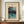 May include: Framed Japanese woodblock print poster showing a winter mountain lake scene with snow-capped peaks, tall trees in foreground, and turquoise water. Print has cream background with museum exhibition text at top and artist signature below. Wooden frame sits on mid-century credenza beside white ceramic vase and stacked books against cream paneled walls.