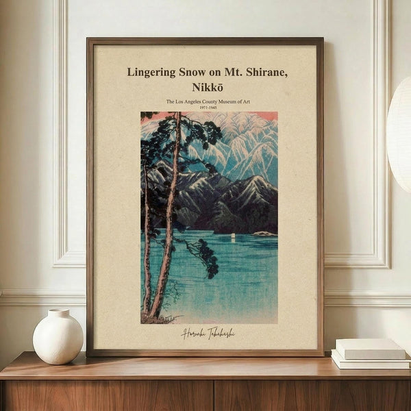 May include: Framed Japanese woodblock print poster showing a winter mountain lake scene with snow-capped peaks, tall trees in foreground, and turquoise water. Print has cream background with museum exhibition text at top and artist signature below. Wooden frame sits on mid-century credenza beside white ceramic vase and stacked books against cream paneled walls.