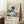May include: Framed poster of Hokusai's famous blue and white Great Wave artwork leaning against white wall next to white coffee cup on wooden block. The vintage-style print shows dramatic ocean wave with foam details and Mount Fuji in background, displayed in light wood frame on white textured rug with beige throw blanket visible.