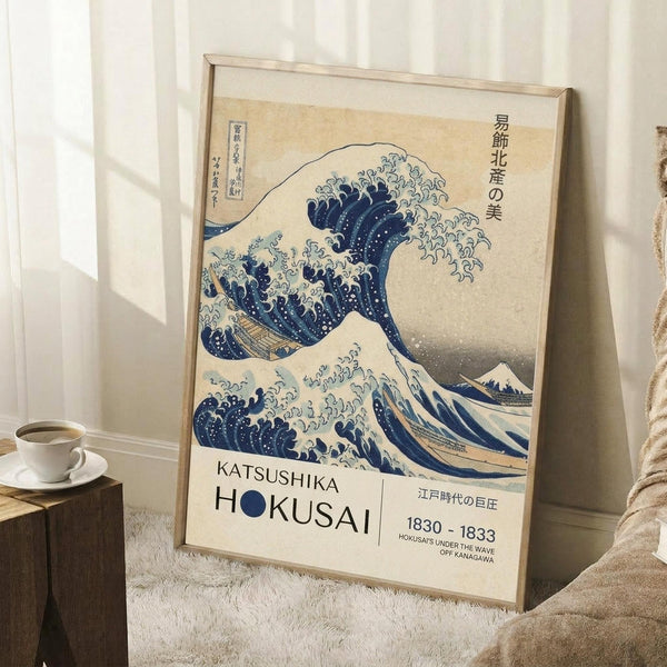 May include: Framed poster of Hokusai's famous blue and white Great Wave artwork leaning against white wall next to white coffee cup on wooden block. The vintage-style print shows dramatic ocean wave with foam details and Mount Fuji in background, displayed in light wood frame on white textured rug with beige throw blanket visible.