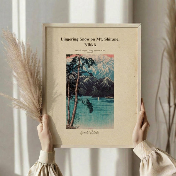 May include: Hands holding a framed Japanese woodblock print showing a serene lake scene with mountains, trees, and turquoise water. The artwork is displayed in a light wooden frame against cream matting. Person wearing beige clothing with gathered sleeves also holds dried wheat stems. Soft natural lighting creates gentle shadows on white curtains in background.
