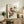 May include: Framed poster featuring two birds with reddish-brown and blue plumage perched on leafy branches, displayed on rustic wooden sideboard alongside fern plant, ceramic vase, and stacked books in neutral-toned living room setting