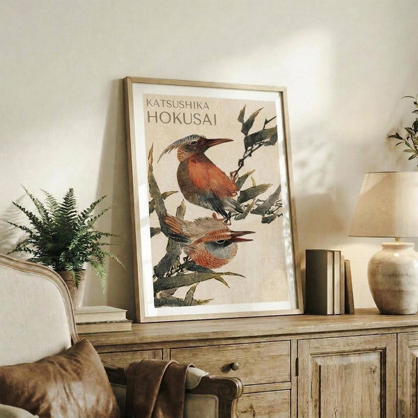 May include: Framed poster featuring two birds with reddish-brown and blue plumage perched on leafy branches, displayed on rustic wooden sideboard alongside fern plant, ceramic vase, and stacked books in neutral-toned living room setting
