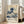 May include: Framed poster of Hokusai's famous blue wave artwork displayed against white wall next to beige upholstered chair. Print shows dramatic ocean wave with white foam in traditional Japanese woodblock style, with museum-style text below including artist name, dates 1830-1833, and Japanese characters. Light wooden frame, natural lighting from windows.