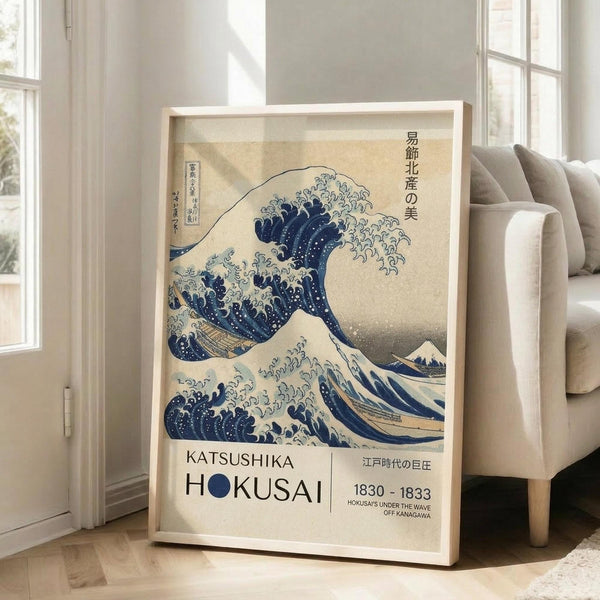 May include: Framed poster of Hokusai's famous blue wave artwork displayed against white wall next to beige upholstered chair. Print shows dramatic ocean wave with white foam in traditional Japanese woodblock style, with museum-style text below including artist name, dates 1830-1833, and Japanese characters. Light wooden frame, natural lighting from windows.