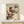 May include: A framed vintage-style Japanese woodblock print featuring two colorful birds perched on green foliage branches against a cream background. The artwork shows detailed feathers in rust, orange, and blue-gray tones with long curved beaks. The print is displayed in a natural wood frame on a light wooden surface alongside a small white wrapped gift with a pine sprig, set against a neutral wall.
