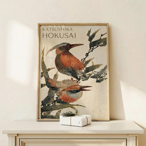 May include: A framed vintage-style Japanese woodblock print featuring two colorful birds perched on green foliage branches against a cream background. The artwork shows detailed feathers in rust, orange, and blue-gray tones with long curved beaks. The print is displayed in a natural wood frame on a light wooden surface alongside a small white wrapped gift with a pine sprig, set against a neutral wall.