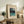 May include: Framed vintage Japanese woodblock print showing snowy mountain landscape with lake and trees, displayed on rustic wooden sideboard alongside potted fern, table lamp, ceramic vase, and stacked books in neutral-toned living room setting with beige upholstered chair visible.