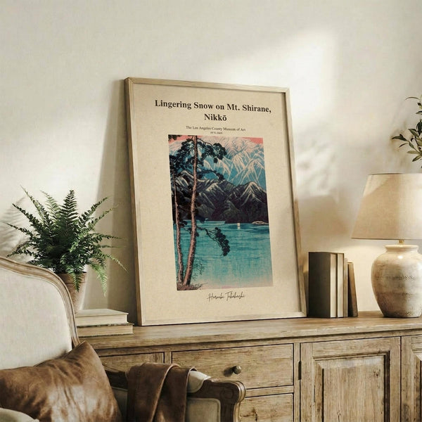 May include: Framed vintage Japanese woodblock print showing snowy mountain landscape with lake and trees, displayed on rustic wooden sideboard alongside potted fern, table lamp, ceramic vase, and stacked books in neutral-toned living room setting with beige upholstered chair visible.