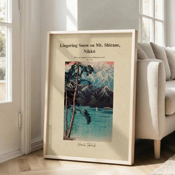 May include: Framed vintage Japanese woodblock print poster showing a serene lake scene with snow-capped mountains, bare trees in foreground, and turquoise water. The artwork features museum-style presentation with cream matting and thin wooden frame, leaning against white wall next to beige upholstered chair in bright, airy room with large windows and natural lighting.