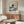 May include: Modern living room with wooden sideboard, white curved chair, glass coffee table, and floor lamp. Wall displays framed poster of famous Japanese wave artwork in blue and cream colors with Mount Fuji in background. Print shows artist name and dates 1830-1831. Minimalist Scandinavian-style decor with warm wood tones and neutral walls.