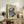 May include: Framed Japanese woodblock print depicting a dramatic waterfall cascading over reddish-brown rocky cliffs with flowing blue and white water, surrounded by green and yellow foliage. The artwork sits on a rustic wooden sideboard next to a fern plant, books, and ceramic vase in a warm, neutral-toned living room setting with a beige upholstered chair visible in the foreground.