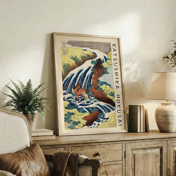 May include: Framed Japanese woodblock print depicting a dramatic waterfall cascading over reddish-brown rocky cliffs with flowing blue and white water, surrounded by green and yellow foliage. The artwork sits on a rustic wooden sideboard next to a fern plant, books, and ceramic vase in a warm, neutral-toned living room setting with a beige upholstered chair visible in the foreground.