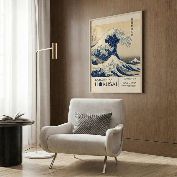 May include: A framed poster of Hokusai's famous blue wave artwork hanging on a wood-paneled wall above a beige upholstered armchair with geometric patterned pillow. A brass floor lamp and dark round side table with open book are positioned nearby. The poster shows the iconic crashing wave in deep blues against a cream background with Japanese text and exhibition details below the image.