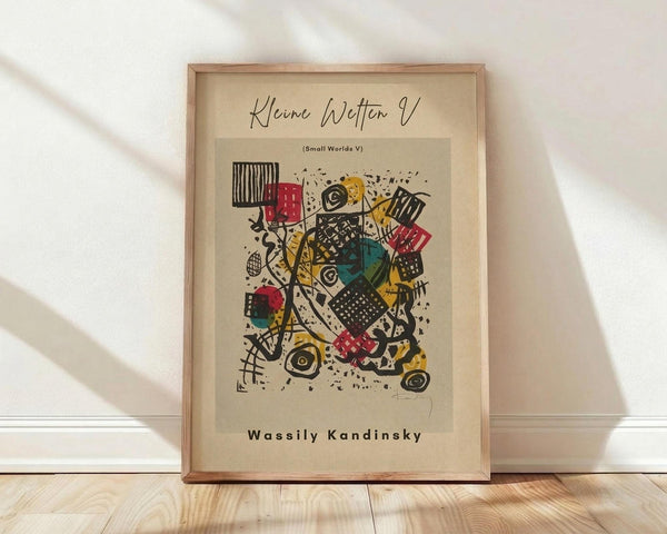 May include: A framed abstract art print by Wassily Kandinsky titled "Small Worlds V" leaning against a white wall on light wooden flooring. The artwork features geometric shapes, lines, and patterns in black, red, yellow, and teal on cream paper. Natural lighting creates soft shadows across the scene. The wooden frame has a natural finish and the print shows Kandinsky's characteristic abstract expressionist style with scattered dots, crosshatched patterns, and dynamic angular forms.