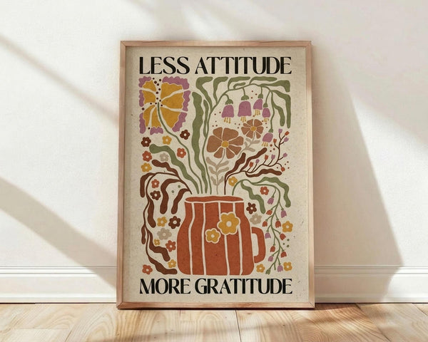 May include: Framed poster with wooden frame leaning against white wall on light wood floor. Features colorful retro-style illustration of potted plant with flowing vines, flowers, and mushrooms in earthy orange, green, purple, and yellow tones. Text reads "Less Attitude More Gratitude" in bold black letters at top and bottom. Artwork has vintage botanical folk art aesthetic on cream background.