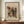 May include: Framed abstract art print in warm wood frame, displayed on wooden sideboard with white ceramic vase and books. The artwork features colorful geometric shapes, patterns and lines in red, yellow, teal and black on cream background. Artist signature visible below the composition. Clean white walls with decorative molding complete the styled room setting.