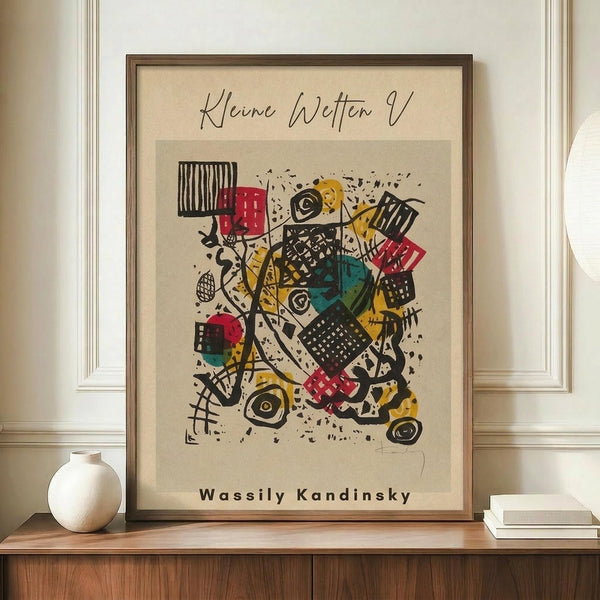 May include: Framed abstract art print in warm wood frame, displayed on wooden sideboard with white ceramic vase and books. The artwork features colorful geometric shapes, patterns and lines in red, yellow, teal and black on cream background. Artist signature visible below the composition. Clean white walls with decorative molding complete the styled room setting.