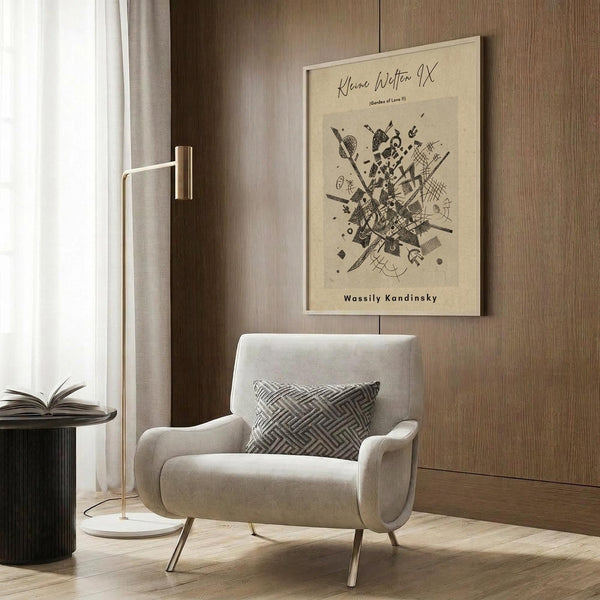 May include: Framed abstract art print with black geometric shapes and lines on cream background, displayed on wood paneled wall above beige upholstered armchair with patterned throw pillow. Modern brass floor lamp and dark round side table with open book visible nearby. Warm neutral toned interior with natural lighting from window.