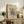 May include: Framed art print with abstract black geometric design on cream background, displayed on rustic wood sideboard beside fern plant, lamp, ceramic vase, and books in neutral-toned living room setting with beige chair visible.