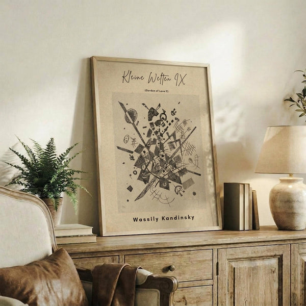 May include: Framed art print with abstract black geometric design on cream background, displayed on rustic wood sideboard beside fern plant, lamp, ceramic vase, and books in neutral-toned living room setting with beige chair visible.