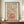May include: Framed wall art print featuring "LESS ATTITUDE MORE GRATITUDE" text with decorative illustration of a beer mug surrounded by flowing botanical elements in warm earth tones of orange, green, brown and pink. The poster has a vintage-inspired design on cream background, displayed in brown wooden frame on wooden sideboard next to white ceramic vase and stacked books in modern room with white paneled walls.