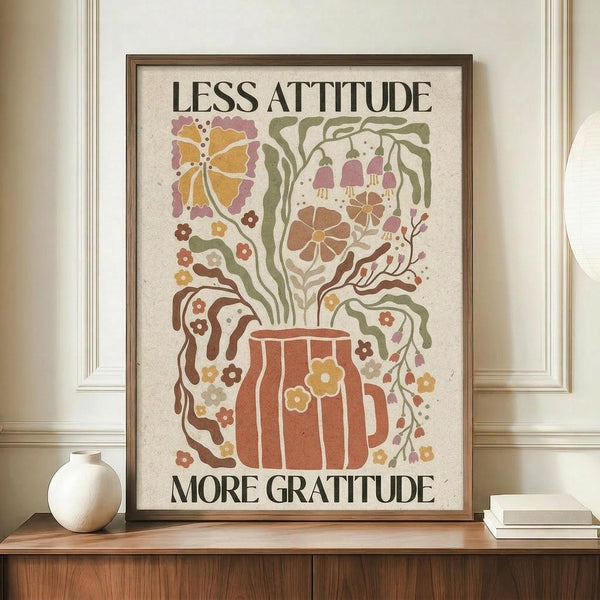 May include: Framed wall art print featuring "LESS ATTITUDE MORE GRATITUDE" text with decorative illustration of a beer mug surrounded by flowing botanical elements in warm earth tones of orange, green, brown and pink. The poster has a vintage-inspired design on cream background, displayed in brown wooden frame on wooden sideboard next to white ceramic vase and stacked books in modern room with white paneled walls.