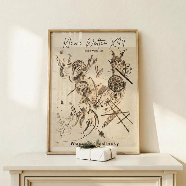 May include: Abstract artwork print in light wooden frame showing dynamic black line drawings and organic shapes on cream background. Framed poster leans against white wall on light wood surface next to small white gift box with greenery sprig. Minimalist modern home decor styling with natural lighting and neutral tones throughout the scene.