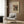 May include: Modern living room with beige upholstered chair and geometric patterned pillow. Framed abstract art poster on wood-paneled wall featuring colorful shapes, patterns and text in red, yellow, green and black. Gold floor lamp with white circular base stands nearby next to dark round side table with open book. Natural lighting from window with sheer curtains illuminates the contemporary space with warm wood flooring.
