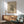 May include: Abstract black geometric artwork with scattered lines, shapes and dots on cream background, displayed in thin wooden frame above mid-century modern wooden credenza. Sage green upholstered armchair with wooden arms sits nearby, white ceramic table lamp on credenza. Warm natural sunlight creates shadows across white wall and wooden furniture in minimalist living room setting.