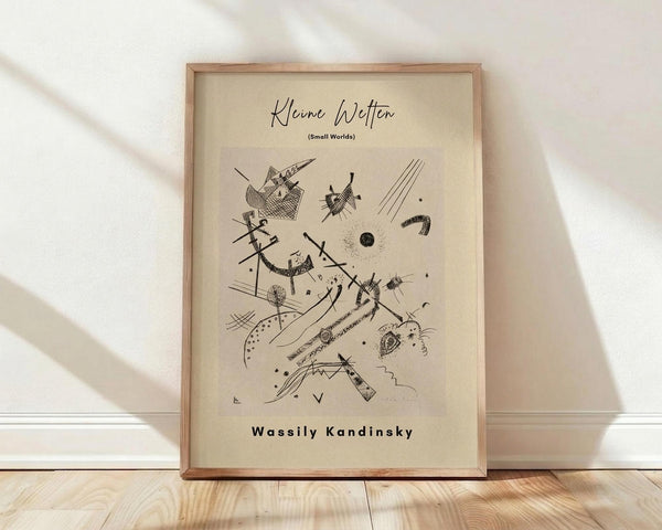 May include: Framed abstract art print by Wassily Kandinsky titled "Kleine Welten (Small Worlds)" featuring black geometric shapes, lines, dots, and angular forms on cream-colored paper. The artwork is displayed in a light wooden frame, leaning against a white wall on light wooden flooring with natural shadows cast across the scene.