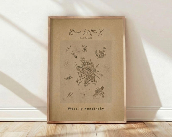 May include: Abstract geometric line drawing poster with beige aged paper texture in wooden frame, featuring scattered angular shapes, stars, and intricate central design. Print displays "Small Worlds X" and "Wassily Kandinsky" text. Frame leans against white wall on light wooden floor with soft natural lighting creating diagonal shadows.
