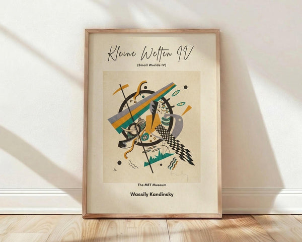 May include: Framed abstract art poster leaning against white wall on light wood floor. Cream-colored print features geometric shapes in orange, teal, black and gray with checkered patterns and organic forms. Thin wooden frame with natural wood grain. Soft natural lighting creates diagonal shadows across white textured wall and smooth wooden planks.