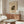 May include: Abstract art poster with geometric shapes in dark green, yellow, and brown tones displayed above a wooden sideboard in a minimalist living room. White curved chair and glass-top coffee table visible in foreground. Poster features Kandinsky-style abstract composition with angular and organic forms on cream background in thin frame.