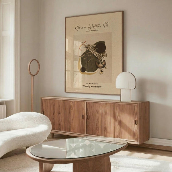 May include: Abstract art poster with geometric shapes in dark green, yellow, and brown tones displayed above a wooden sideboard in a minimalist living room. White curved chair and glass-top coffee table visible in foreground. Poster features Kandinsky-style abstract composition with angular and organic forms on cream background in thin frame.
