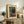 May include: Framed vintage Japanese woodblock print showing a moonlit lake scene with dark silhouetted trees, mountains, and traditional buildings reflected in blue water. The artwork is displayed on a rustic wooden sideboard alongside books, a ceramic vase, and a potted fern, with a cream-colored armchair visible in a warm, neutral-toned living room setting.