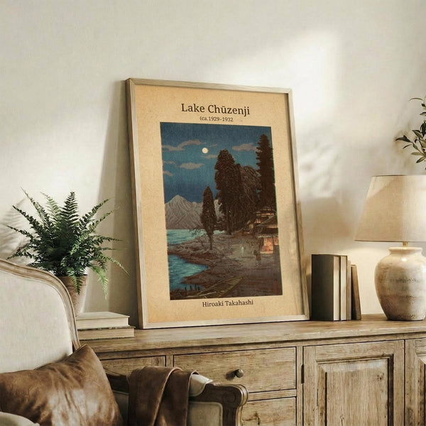 May include: Framed vintage Japanese woodblock print showing a moonlit lake scene with dark silhouetted trees, mountains, and traditional buildings reflected in blue water. The artwork is displayed on a rustic wooden sideboard alongside books, a ceramic vase, and a potted fern, with a cream-colored armchair visible in a warm, neutral-toned living room setting.