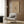 May include: Modern reading nook with cream upholstered armchair featuring geometric throw pillow, brass floor lamp with white circular base, dark round side table with open book, and framed beige art print showing abstract linear drawings mounted on wood-paneled wall. Warm neutral tones throughout with natural lighting from window.