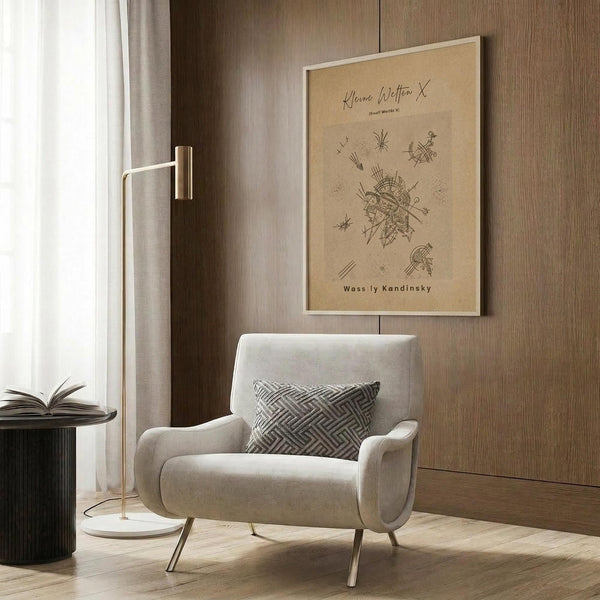 May include: Modern reading nook with cream upholstered armchair featuring geometric throw pillow, brass floor lamp with white circular base, dark round side table with open book, and framed beige art print showing abstract linear drawings mounted on wood-paneled wall. Warm neutral tones throughout with natural lighting from window.