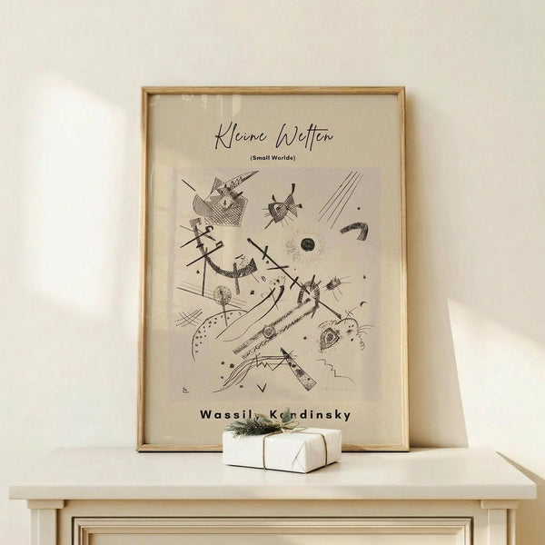May include: Framed abstract art print featuring black geometric shapes, lines, dots and organic forms on cream background, displayed on light wood console table with small white wrapped gift box beside it, against neutral wall in minimalist room setting
