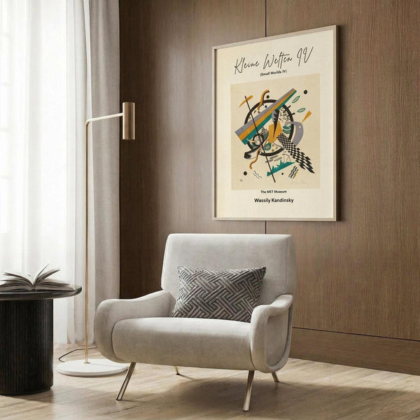 May include: Abstract Kandinsky poster with geometric shapes in orange, teal, black and beige, displayed in white frame on wood paneled wall above beige upholstered chair with geometric patterned pillow. Modern brass floor lamp and dark round side table with open book complete the warm, contemporary room setting with wood floors and white curtains.