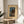 May include: Framed vintage Japanese woodblock print displayed on round wooden side table next to beige upholstered chair. Artwork shows moonlit lake scene with traditional buildings, tall cypress trees, and mountain backdrop in blue and earth tones. Natural lighting creates soft shadows across cream-colored matting and thin wooden frame.