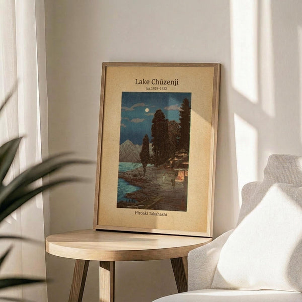 May include: Framed vintage Japanese woodblock print displayed on round wooden side table next to beige upholstered chair. Artwork shows moonlit lake scene with traditional buildings, tall cypress trees, and mountain backdrop in blue and earth tones. Natural lighting creates soft shadows across cream-colored matting and thin wooden frame.