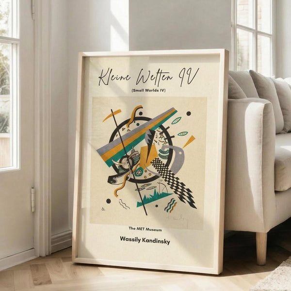 May include: Framed abstract art print leaning against white wall next to beige upholstered chair in bright sunlit room with large windows. The poster features Kandinsky's geometric composition with intersecting black lines, orange and teal diagonal bands, checkered patterns, and small decorative elements on cream background. Natural wood frame, approximately 2-3 feet tall, casting soft shadows on light wooden floor.