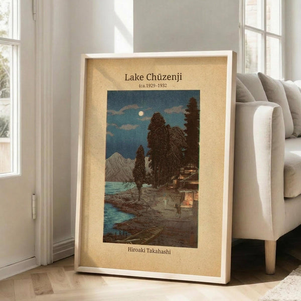 May include: Framed vintage Japanese woodblock print showing a moonlit lakeside scene with traditional buildings, tall trees, mountains, and blue water. The artwork has a cream-colored mat border and white frame, leaning against a white wall next to a beige sectional sofa in a bright living room with hardwood floors and large windows.