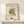 May include: Framed abstract art print in light wood frame displaying geometric shapes, lines, and figures in orange, teal, black, and gray on cream background. Print sits on pale wooden surface against white wall next to small white gift box with greenery sprig.
